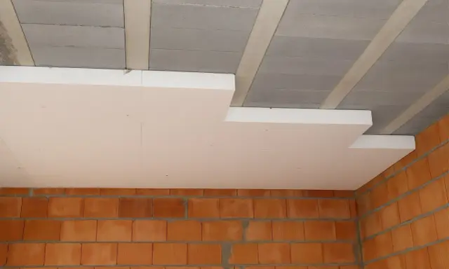 Ocieplenie stropu styropianem w trakcie budowy. Białe płyty styropianowe montowane do betonowych belek stropowych, widoczne ceglane ściany.