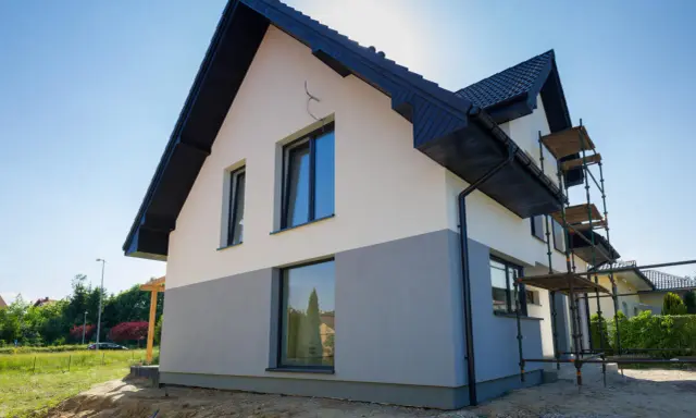 Nowy dom w trakcie budowy, z dwukolorową elewacją i rusztowaniami. Czekamy na wykończenie, by podziwiać efekt końcowy.